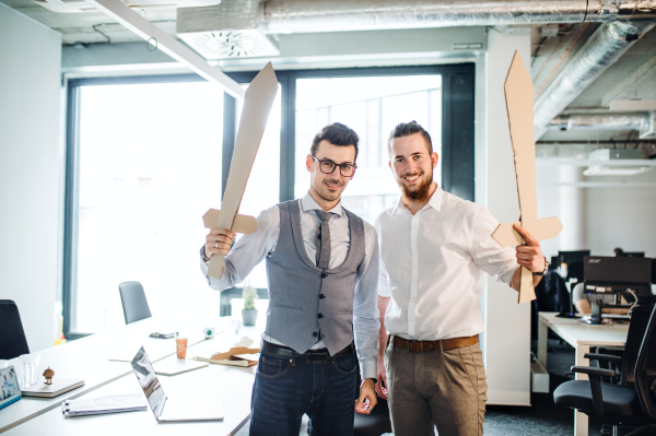 Two young businessmen with cardboard swords in an office. A competition concept.