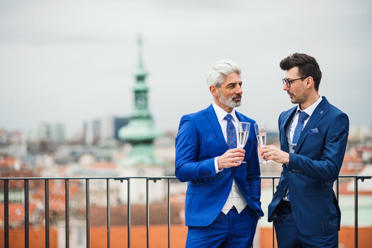 Two businessmen with champagne standing outdoors on roof terrace in city.