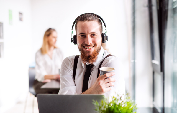 A portrait of young businessman with laptop, headphones and coffee in an office, looking at camera.