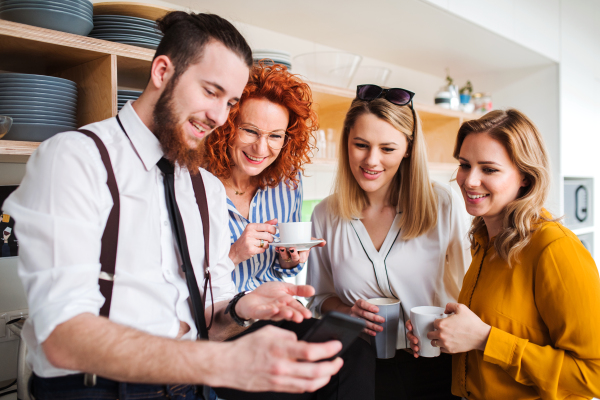 A group of young cheerful business people on coffee break in office kitchen, taking selfie.