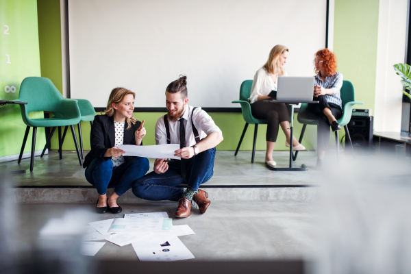 A group of young business people with a diary sitting on the floor in an office, talking.