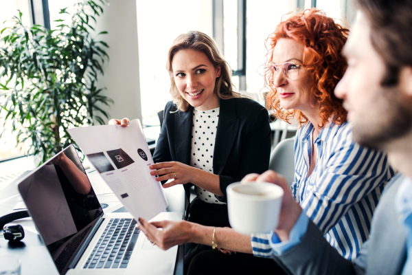 A group of young cheerful business people with laptop sitting in an office, talking.