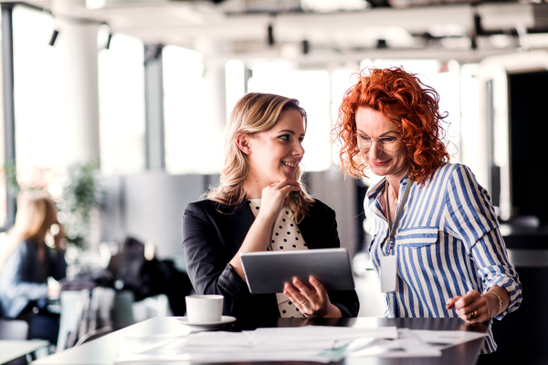 Two cheerful businesswomen with tablet sitting in an office, talking.