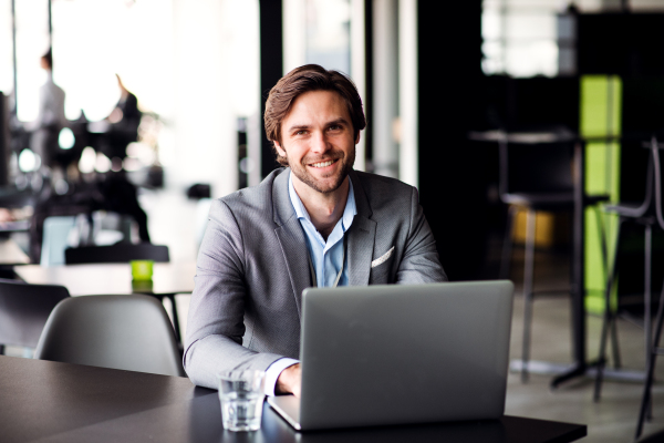 A portrait of happy young businessman with computer in an office, looking at camera.