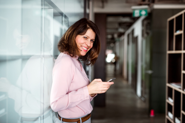 A portrait of businesswoman with smartphone standing in an office building, text messaging.