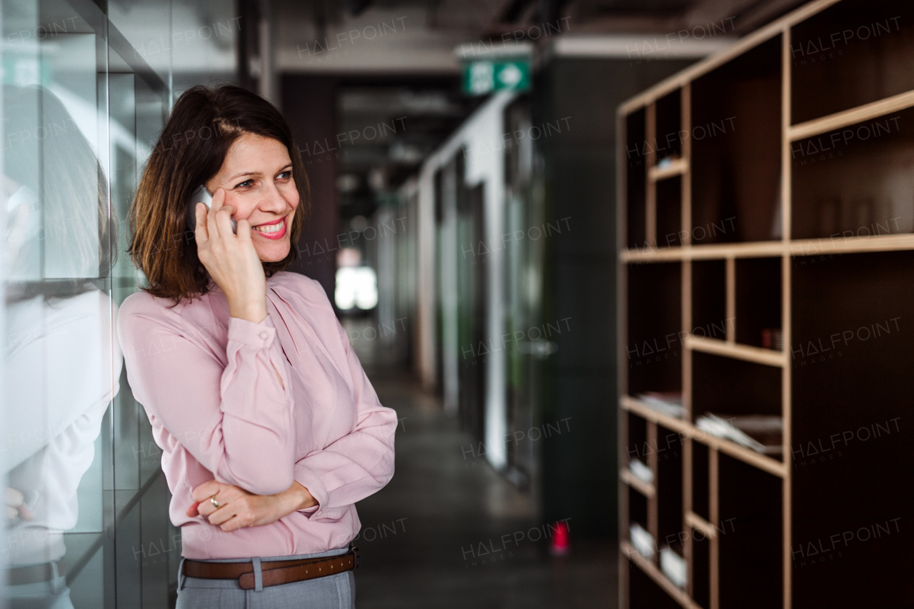 A portrait of businesswoman with smartphone standing in an office building with crossed arms, making a phone call.