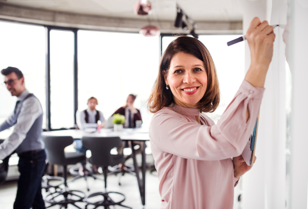 A portrait of businesswoman with colleagues standing in an office, writing on whiteboard.