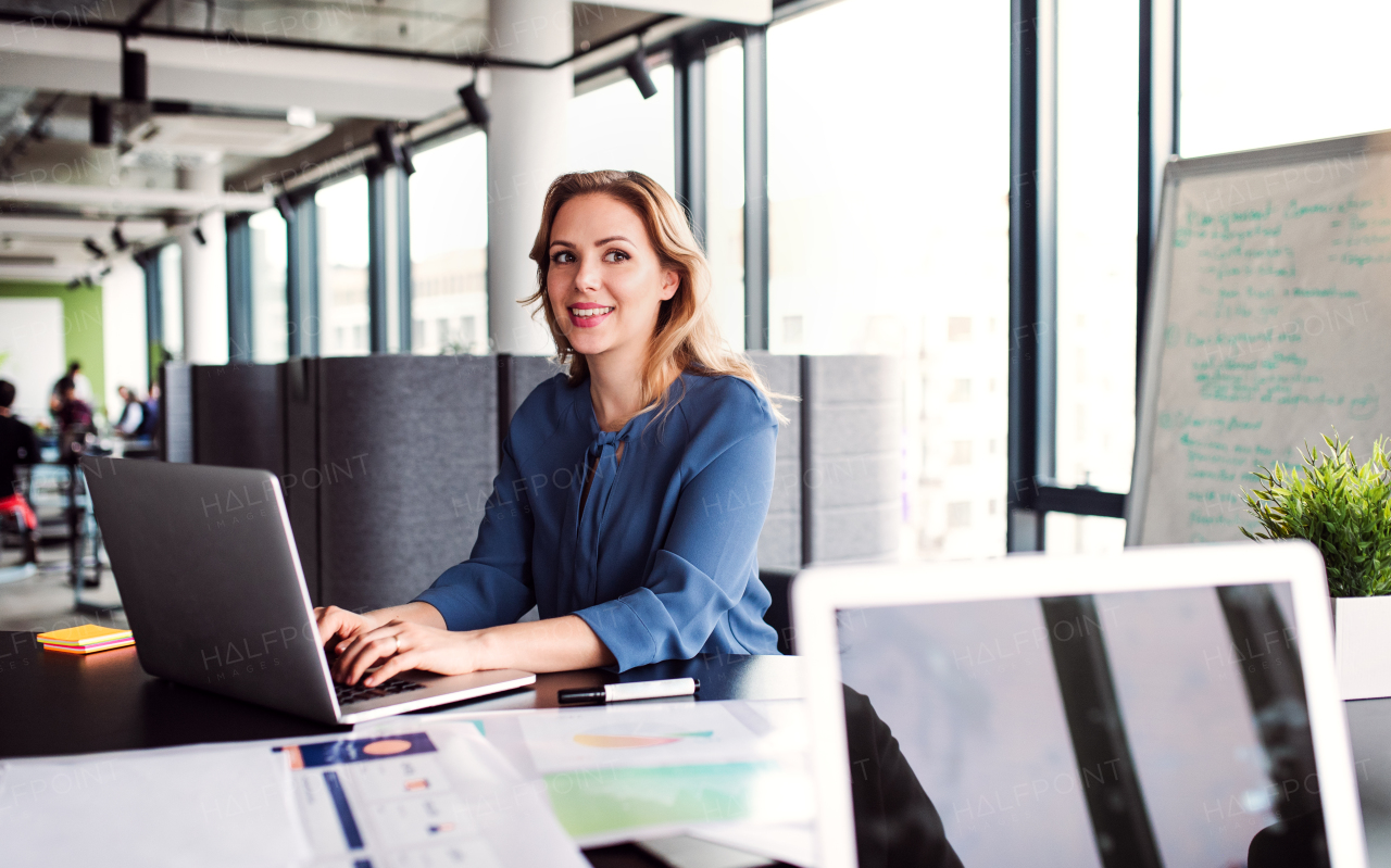 Young beautiful businesswoman with laptop sitting in an office, looking at camera.