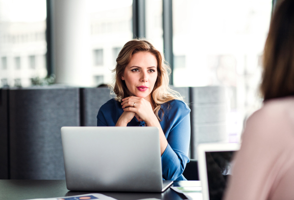 Young beautiful businesswoman with laptop sitting in an office, talking to a colleague.