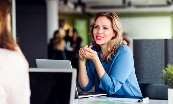 Young beautiful businesswoman with laptop sitting in an office, talking to a colleague.