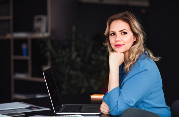 Young beautiful businesswoman with laptop sitting in an office, looking at camera.