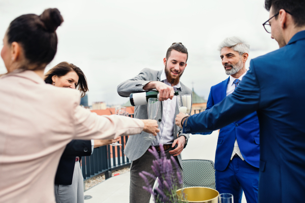 A large group of joyful businesspeople having a party outdoors on roof terrace in city, pouring champagne.