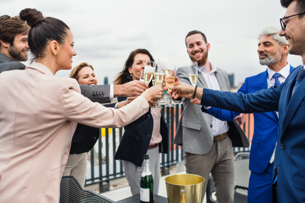 A large group of joyful businesspeople having a party outdoors on roof terrace in city, clinking glasses.