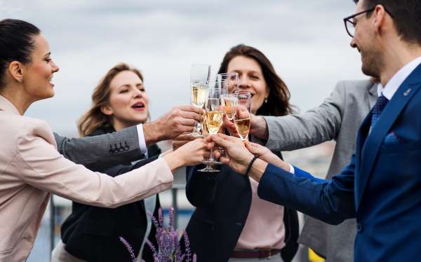 A large group of joyful businesspeople having a party outdoors on roof terrace in city, clinking glasses.