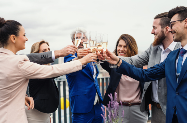 A large group of joyful businesspeople having a party outdoors on roof terrace in city, clinking glasses.