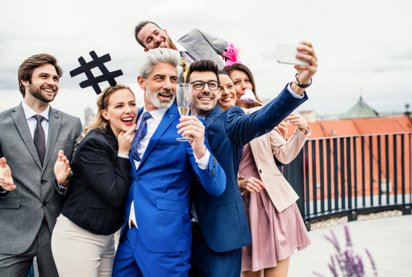 A large group of joyful businesspeople having a party outdoors on roof terrace in city, taking selfie.