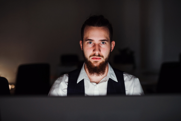 A portrait of young hipster businessman with computer in an office, looking at camera.