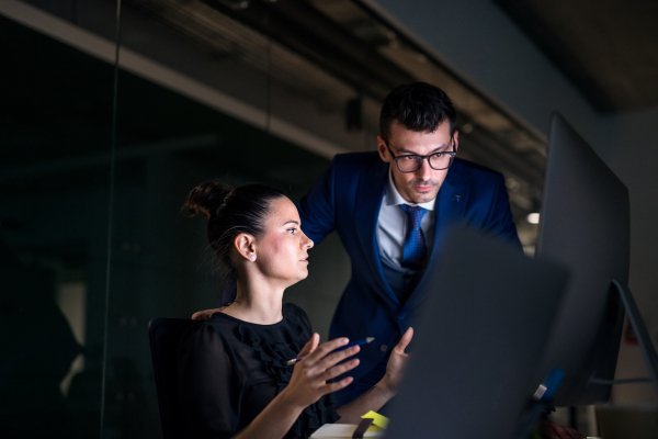 Two young business people in an office in the evening or at night, using computer.