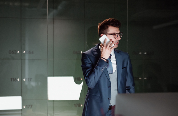 A portrait of young businessman with smartphone standing by lockers in an office, making a phone call. Copy space.