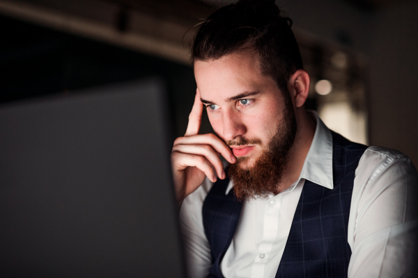 A portrait of young thoughtful hipster businessman with computer in an office, working.