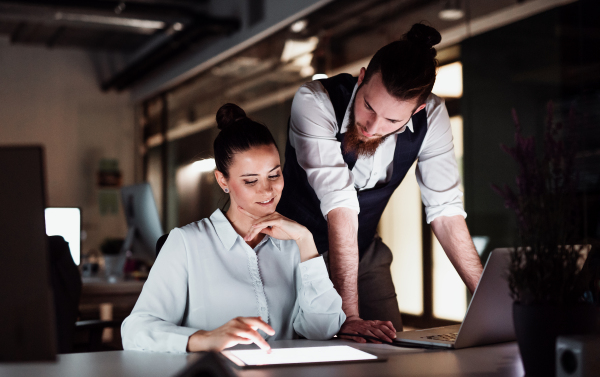 Two young business people in an office in the evening or at night, using computer.