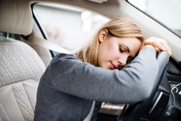 A tired young woman driver sitting in car, head resting on steering wheel.