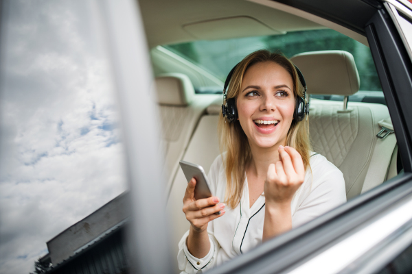 Business woman with smartphone and headphones sitting on back seats in taxi car, listening to music.