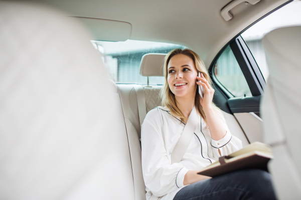 Business woman with smartphone and laptop sitting on back seats in taxi car, talking on the phone.