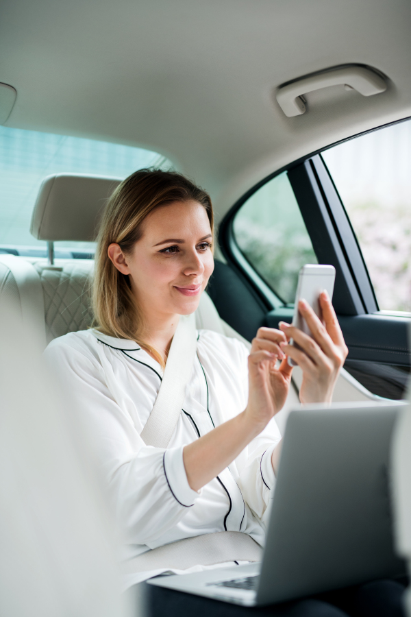 A businesswoman with laptop sitting on back seat in taxi car, taking selfie.