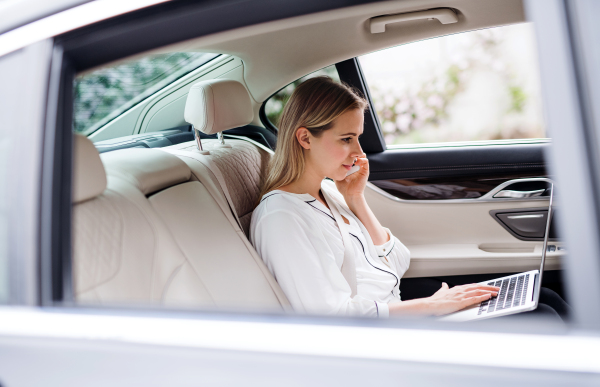 Business woman with smartphone and laptop sitting on back seats in taxi car, talking on the phone.