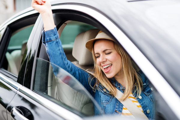 A young cheerful woman with hat sitting in car, listening to music and dancing.