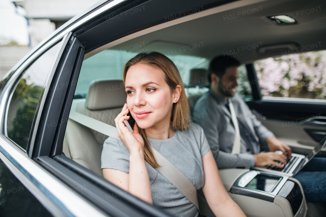 Young business couple with laptop and smartphone sitting on back seats in car, working.