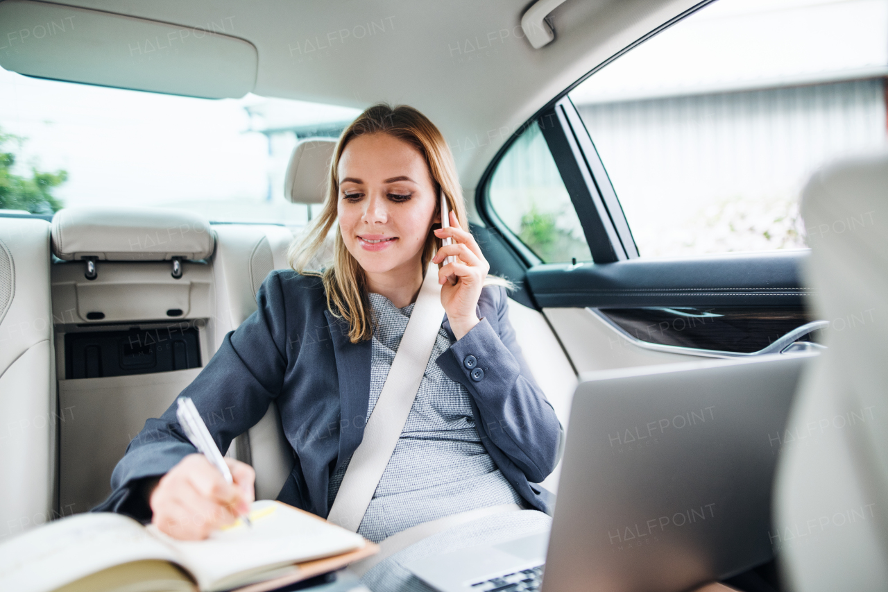 Business woman with smartphone and laptop sitting on back seats in taxi car, talking on the phone.