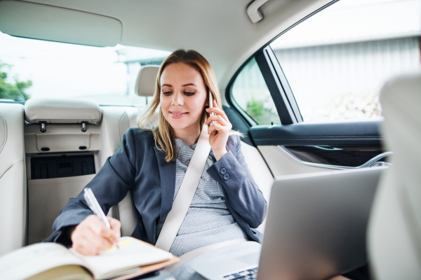 Business woman with smartphone and laptop sitting on back seats in taxi car, talking on the phone.