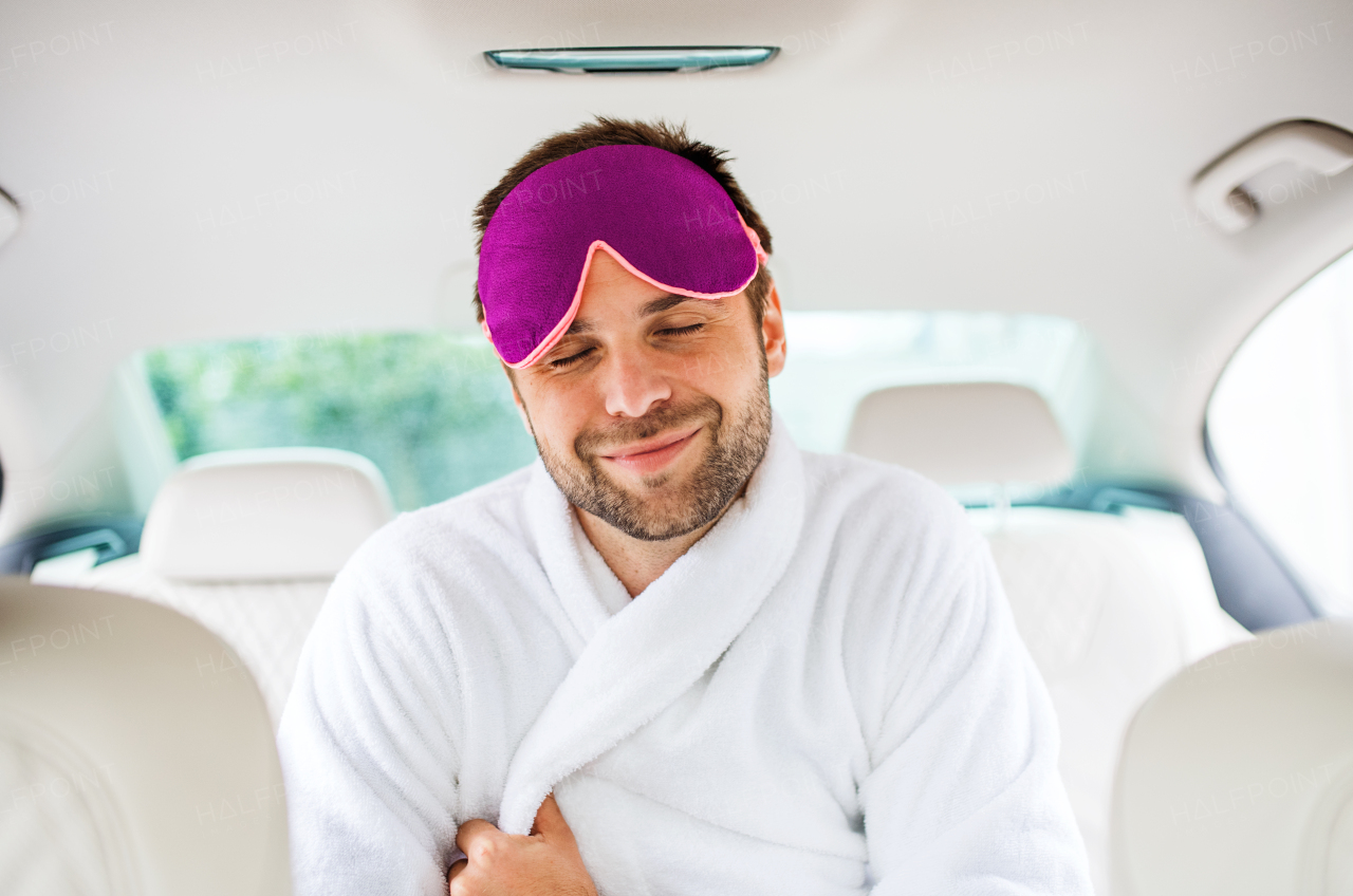 A cheerful man with dressing gown and eye mas sitting in car, eyes closed.