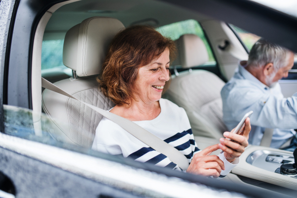 A happy senior couple with smartphone sitting in car, going on trip.