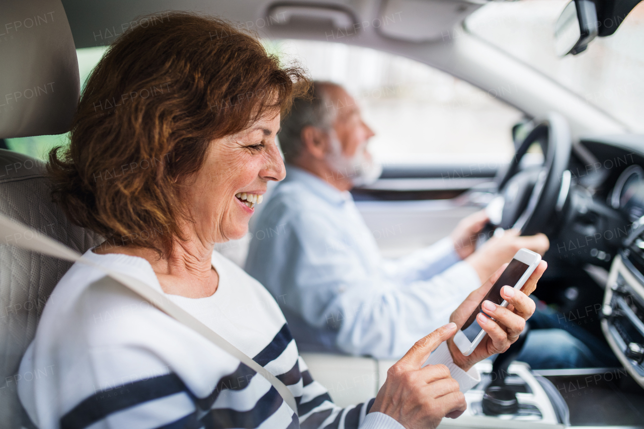 A happy senior couple with smartphone sitting in car, going on trip.