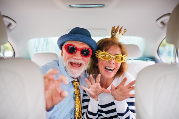 A cheerful senior couple with party accessories sitting in car, having fun.