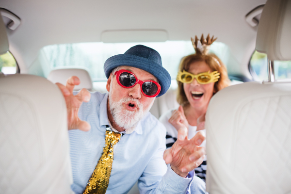 A cheerful senior couple with party accessories sitting in car, having fun.