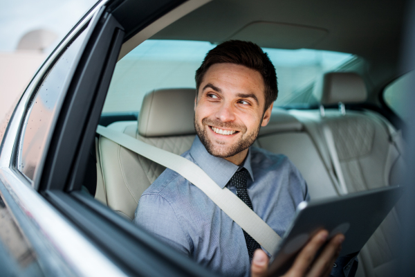 A businessman with tablet sitting on back seats in car, working.