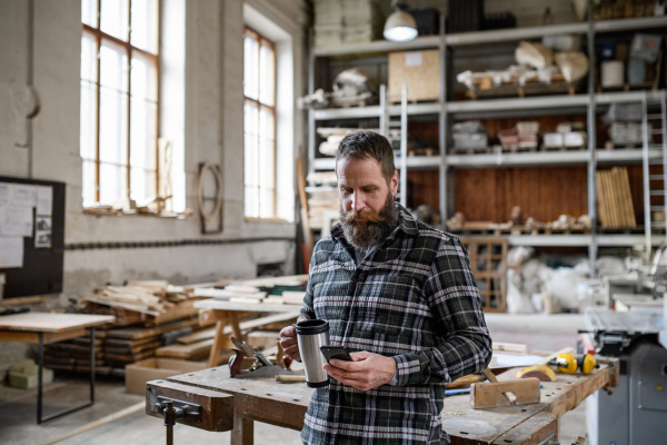 A portrait of mature male carpenter with coffee and smrtphone indoors in carpentery workshop.