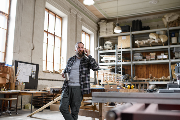 A happy mature male carpenter making phone call during coffee break indoors in carpentery workshop.