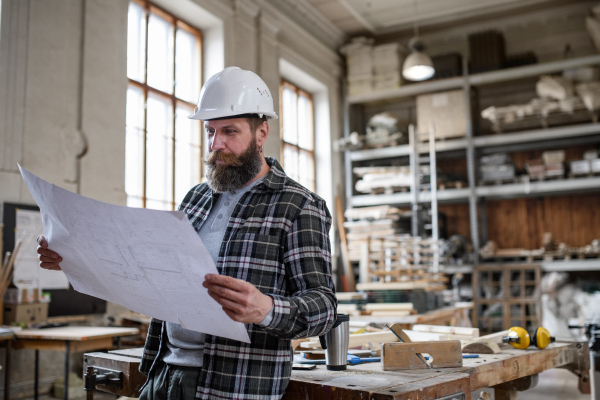 A mature male carpenter looking at blueprints plans in carpentery workshop. Small business concept.