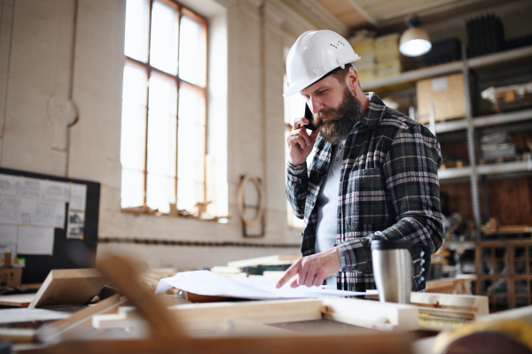 A portrait of mature male carpenter looking at blueprints plans and making a phone call in carpentery workshop. Small business concept.