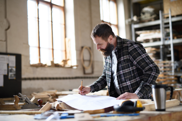 Low angle view of a mature male carpenter looking at blueprints plans in carpentery workshop. Small business concept.