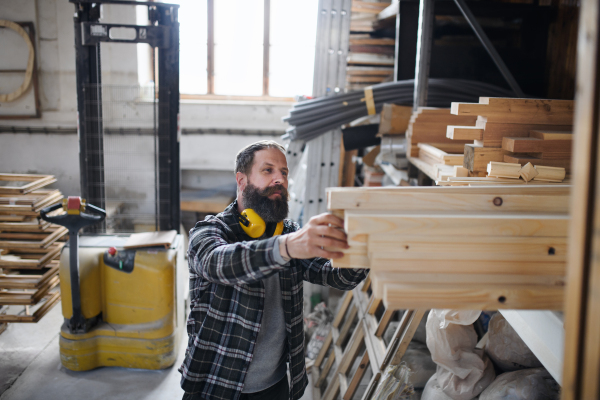 Mature male carpenter with a wooden board indoors in carpentery workshop. Small business concept.