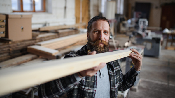 A mature male carpenter carrying wooden board indoors in carpentery workshop. Small business concept.