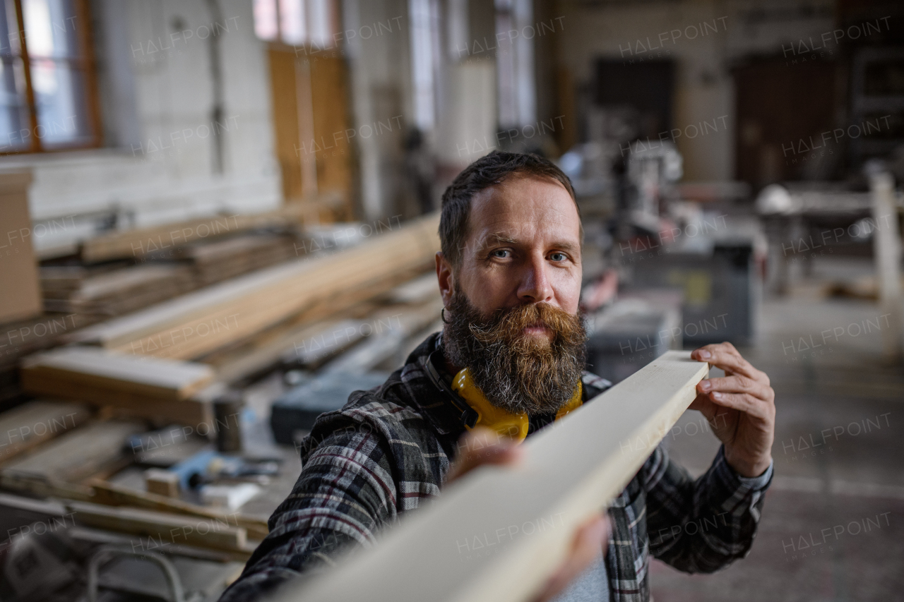 A mature male carpenter carrying wooden board indoors in carpentery workshop. Small business concept.