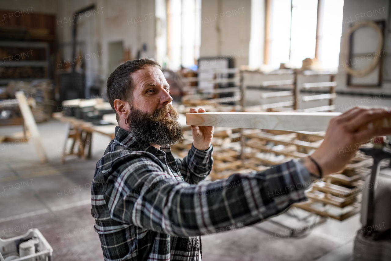 A mature male carpenter carrying wooden board indoors in carpentery workshop. Small business concept.