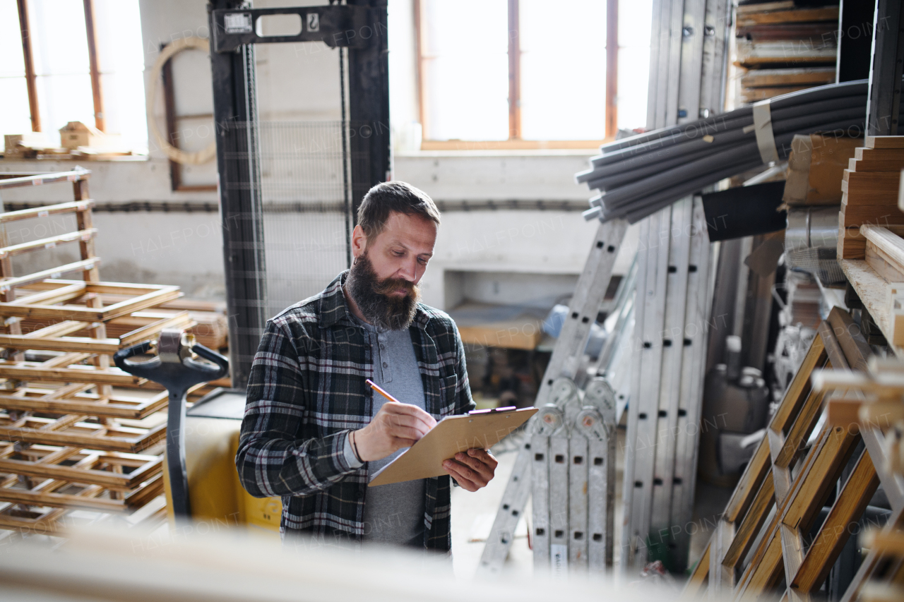 Mature male carpenter doing an inventory indoors in carpentery workshop. Small business concept.